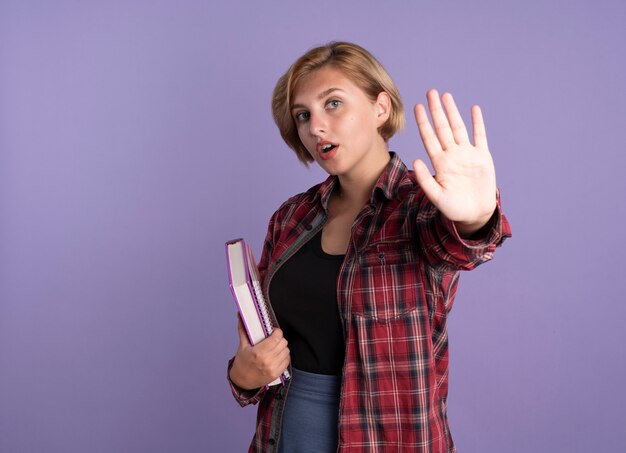 Chica joven estudiante eslava confiada sostiene gestos de libro y cuaderno dejar señal de mano