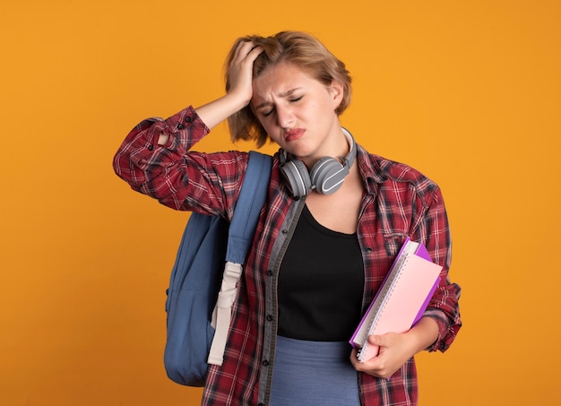 Foto gratuita chica joven estudiante eslava dolorida con auriculares con mochila pone la mano en la frente sostiene el libro y el cuaderno