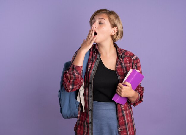 Chica joven estudiante eslava soñolienta con mochila pone la mano en la boca sostiene el libro y el cuaderno