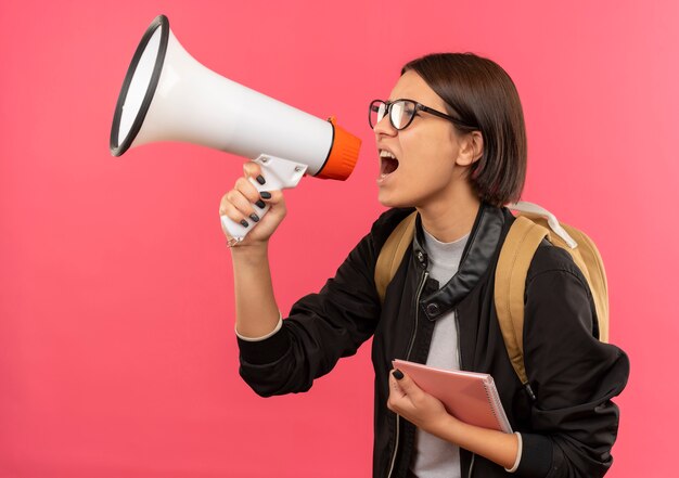 Chica joven estudiante con gafas y bolsa trasera de pie en la vista de perfil sosteniendo el bloc de notas hablando por altavoz aislado sobre fondo rosa