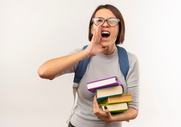 Chica joven estudiante con gafas y bolsa trasera sosteniendo libros poniendo la mano cerca de la boca llamando a alguien aislado sobre fondo blanco.