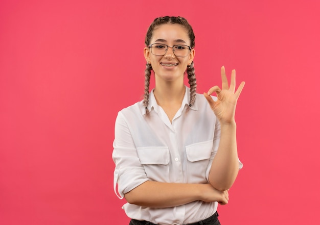 Foto gratuita chica joven estudiante en gafas con coletas en camisa blanca mirando hacia el frente con una sonrisa en la cara mostrando el signo de ok de pie sobre la pared rosa