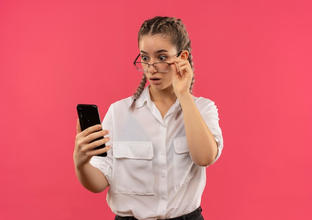 Chica joven estudiante en gafas con coletas en camisa blanca mirando la pantalla de su teléfono móvil asombrado y sorprendido de pie sobre la pared rosa