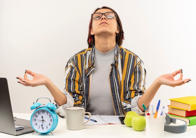 Foto gratuita chica joven estudiante pacífica con gafas sentado en el escritorio meditando con los ojos cerrados aislado sobre fondo blanco.