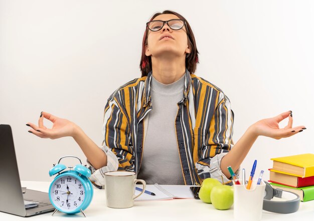 Chica joven estudiante pacífica con gafas sentado en el escritorio meditando con los ojos cerrados aislado sobre fondo blanco.