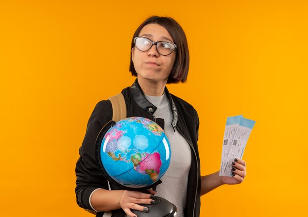 Chica joven estudiante pensativa con gafas y bolsa trasera con globo y entradas mirando al lado aislado en la pared naranja