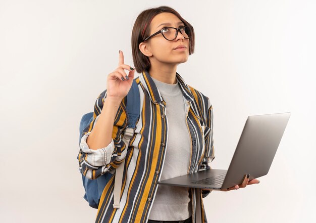 Chica joven estudiante pensativa con gafas y bolsa trasera sosteniendo portátil mirando y apuntando hacia arriba aislado en la pared blanca