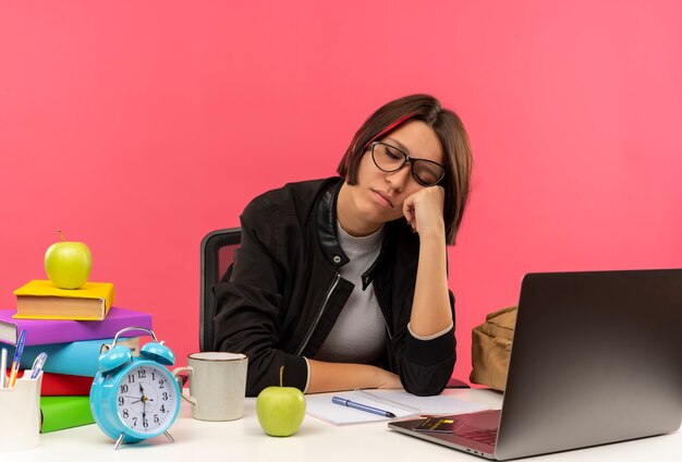 Chica joven estudiante soñolienta con gafas sentado en el escritorio poniendo la mano en la mejilla con los ojos cerrados aislado en la pared rosa