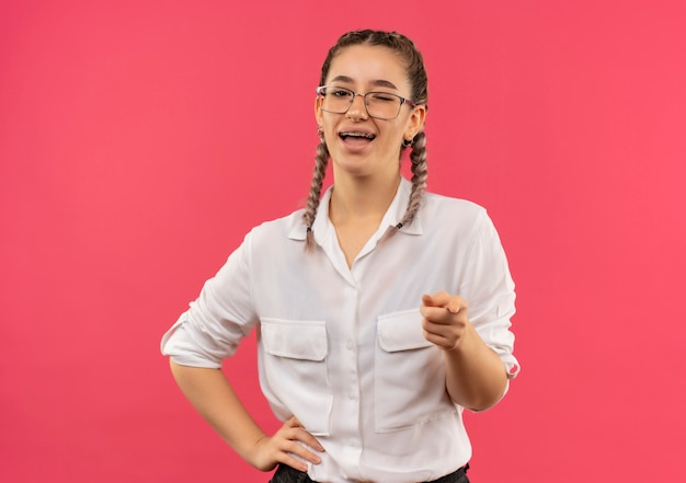 Chica joven estudiante en vasos con coletas en camisa blanca apuntando con el dedo índice al frente sonriendo ampliamente feliz y positivo de pie sobre la pared rosa