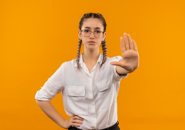 Chica joven estudiante en vasos con coletas en camisa blanca haciendo señal de stop con la mano mirando hacia el frente con cara seria de pie sobre la pared naranja