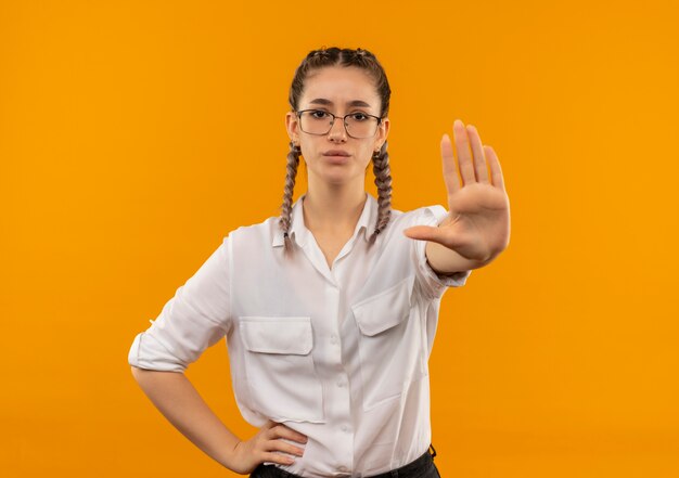 Chica joven estudiante en vasos con coletas en camisa blanca haciendo señal de stop con la mano mirando hacia el frente con cara seria de pie sobre la pared naranja