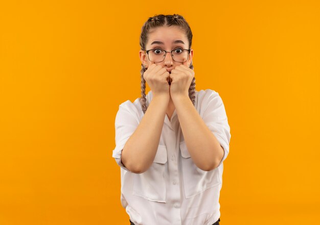 Chica joven estudiante en vasos con coletas en camisa blanca mirando al frente estresado y preocupado morderse las uñas de pie sobre la pared naranja