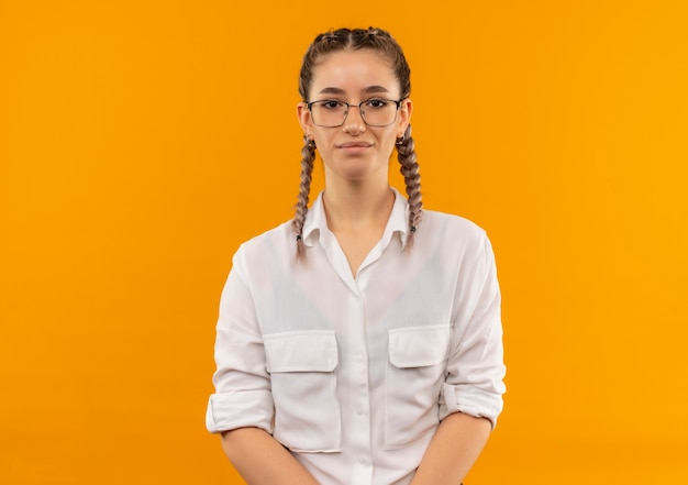 Chica joven estudiante en vasos con coletas en camisa blanca mirando al frente con expresión seria y segura de pie sobre la pared naranja