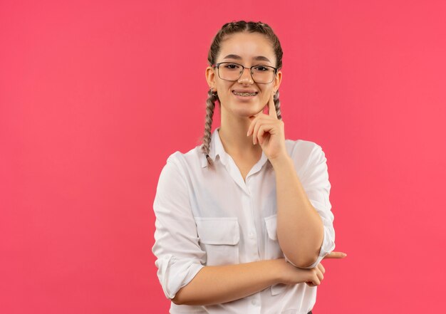 Chica joven estudiante en vasos con coletas en camisa blanca mirando al frente sonriendo confiado de pie sobre la pared rosa