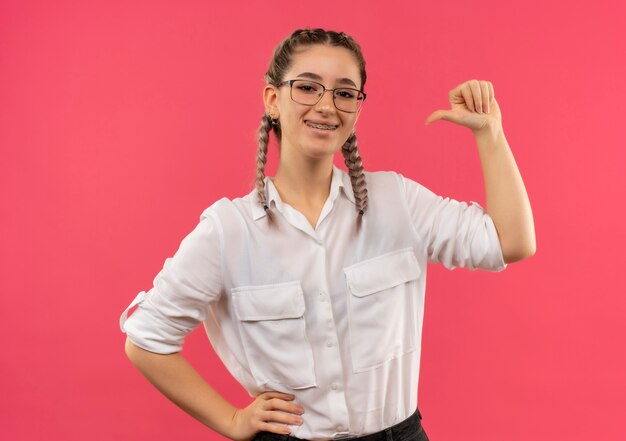 Chica joven estudiante en vasos con coletas en camisa blanca mirando al frente sonriendo feliz y positivo apuntando a sí misma de pie sobre la pared rosa