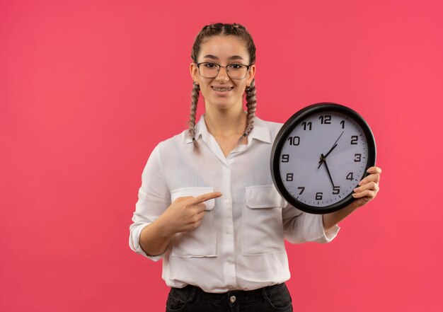 Foto gratuita chica joven estudiante en vasos con coletas en camisa blanca con reloj de pared apuntando con el dedo hacia él, mirando al frente sonriendo confiado de pie sobre la pared rosa