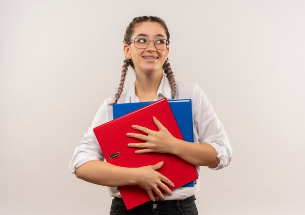 Chica joven estudiante en vasos con coletas en camisa blanca sosteniendo carpetas mirando a un lado con una sonrisa en la cara de pie sobre la pared blanca