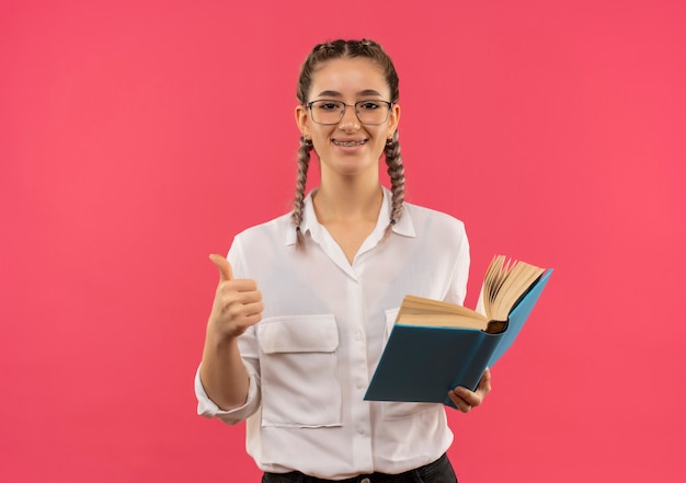 Chica joven estudiante en vasos con coletas en camisa blanca sosteniendo un libro abierto mirando al frente sonriendo mostrando los pulgares para arriba de pie sobre la pared rosa