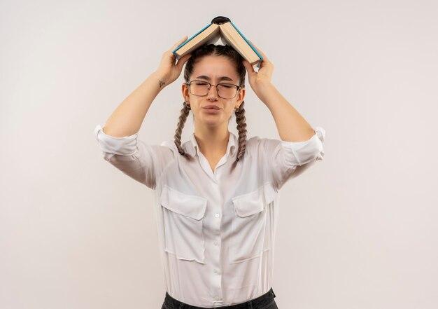 Chica joven estudiante en vasos con coletas en camisa blanca sosteniendo un libro abierto sobre su cabeza mirando decepcionado de pie sobre la pared blanca