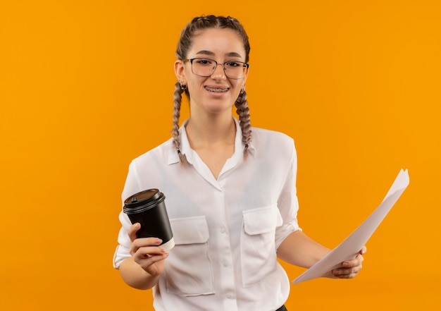 Chica joven estudiante en vasos con coletas en camisa blanca con taza de café y páginas en blanco mirando al frente sonriendo confiado de pie sobre la pared naranja