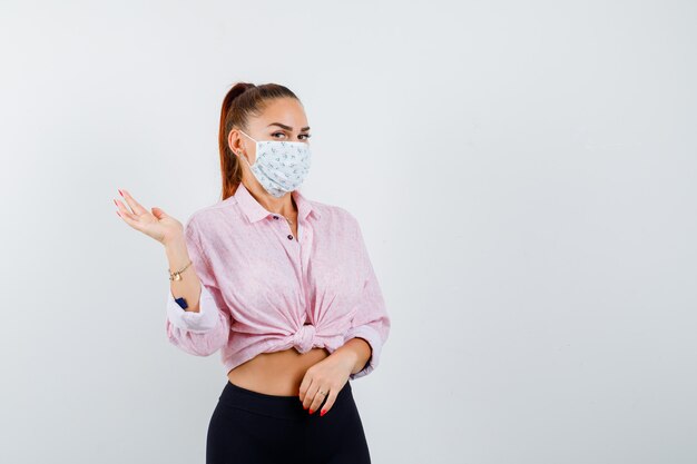 Chica joven extendiendo la palma a un lado en blusa rosa, pantalón negro, máscara y bonita vista frontal.