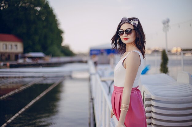 Chica joven con falda rosa y gafas de sol posando en una barandilla