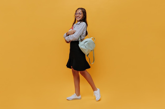 Chica joven feliz en falda negra y camisa sostiene cuadernos y mochila sobre fondo naranja Alegre adolescente sonríe en aislado