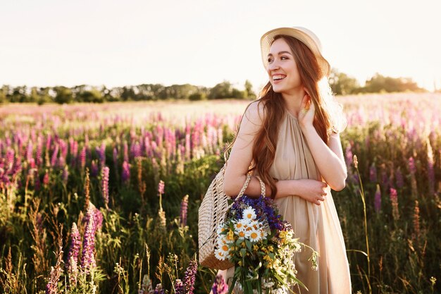 Chica joven feliz que camina en campo de flor en la puesta del sol. Llevaba sombrero de paja y bolso lleno de flores.