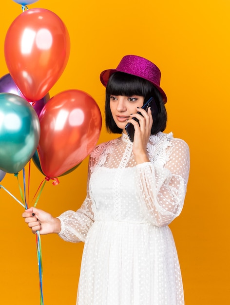 Chica joven fiestera con gorro de fiesta sosteniendo globos hablando por teléfono mirando al lado aislado en la pared naranja