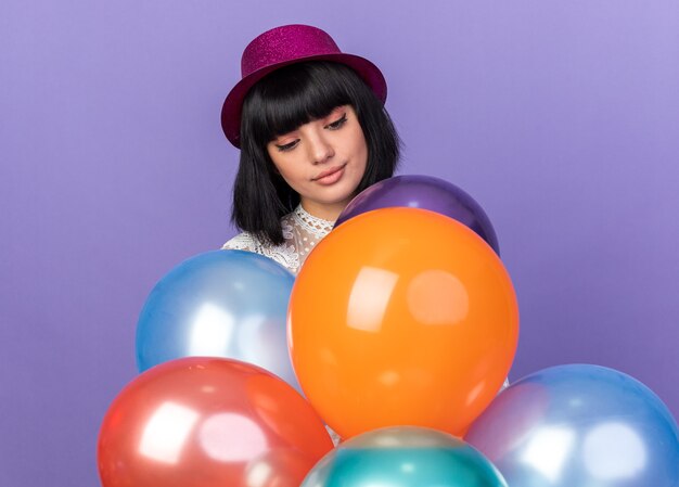 Chica joven fiestera con sombrero de fiesta de pie detrás de globos mirándolos aislado en la pared púrpura