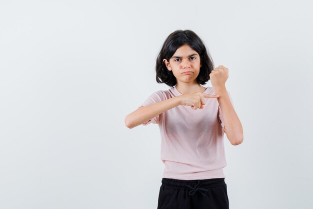 Chica joven fingiendo como señalar el reloj en camiseta rosa y pantalón negro y luciendo linda