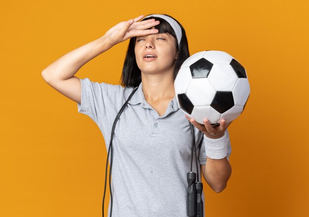 Chica joven fitness con diadema con cuerda alrededor del cuello sosteniendo un balón de fútbol con aspecto cansado y con exceso de trabajo con la mano en la frente de pie sobre fondo naranja