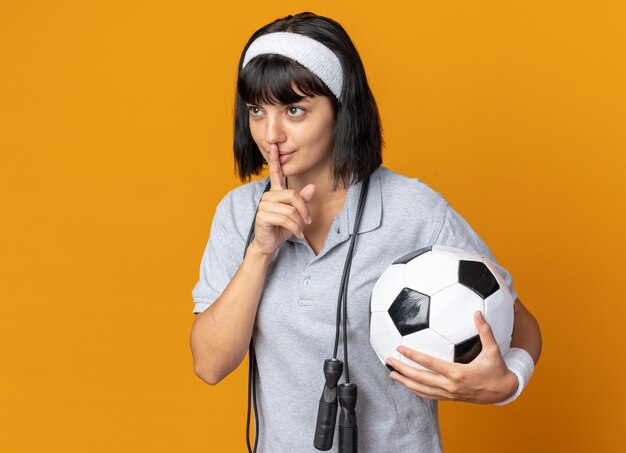 Chica joven fitness con diadema con cuerda alrededor del cuello sosteniendo un balón de fútbol haciendo gesto de silencio con el dedo en los labios de pie sobre fondo naranja