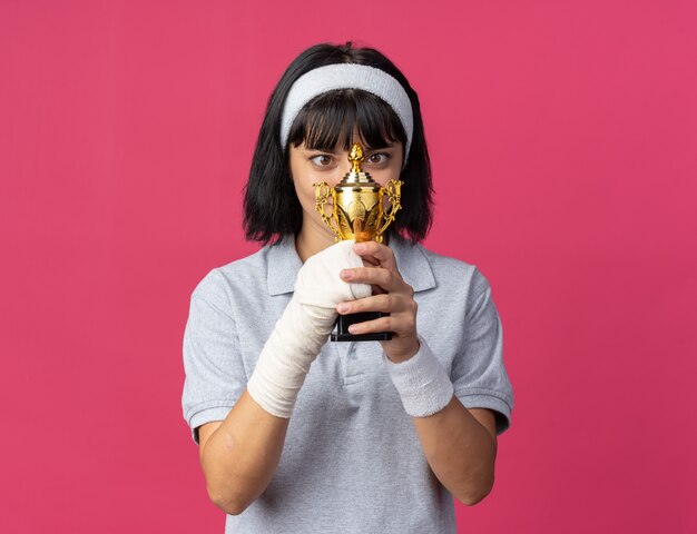 Chica joven fitness con diadema con mano vendada sosteniendo el trofeo mirándolo sorprendido y feliz