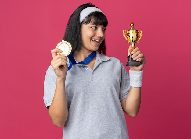 Chica joven fitness con diadema con medalla de oro alrededor del cuello sosteniendo el trofeo mirándolo feliz y emocionado de pie sobre rosa