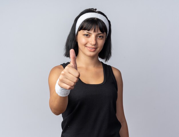 Chica joven fitness con diadema mirando a la cámara sonriendo seguros mostrando el pulgar hacia arriba de pie sobre blanco