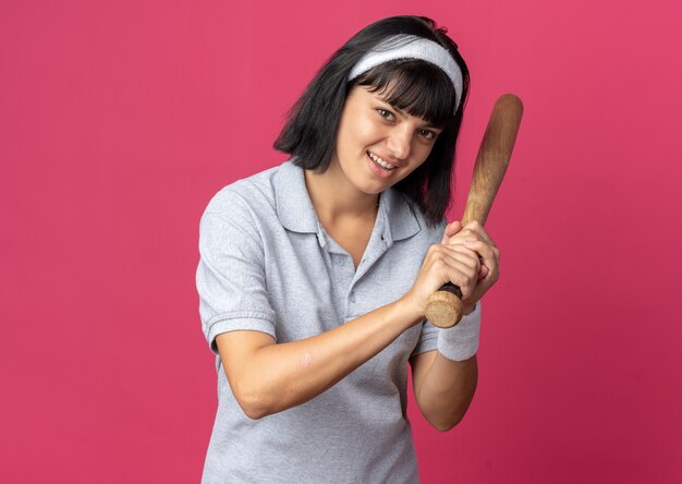 Chica joven fitness con diadema sosteniendo un bate de béisbol mirando a la cámara sonriendo feliz y positivo de pie sobre fondo de color rosa