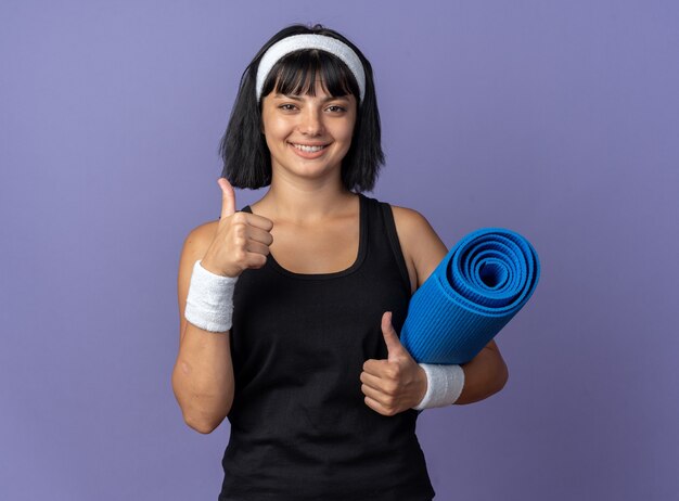 Chica joven fitness con diadema sosteniendo una estera de yoga mirando a la cámara sonriendo alegremente mostrando los pulgares para arriba de pie sobre fondo azul.