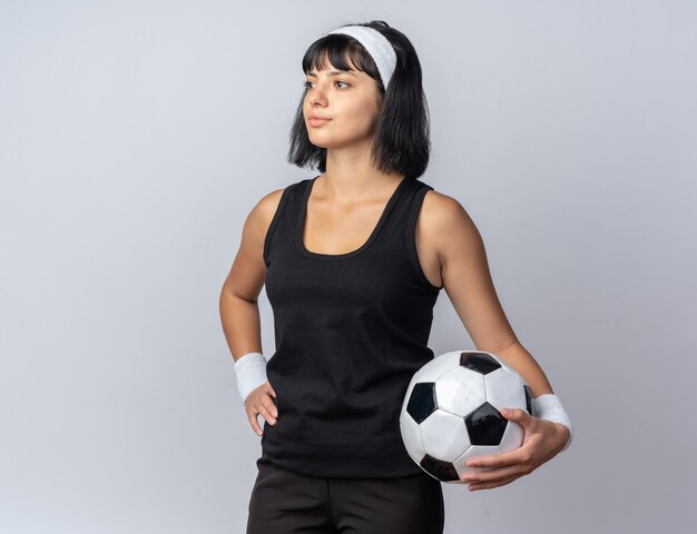 Chica joven fitness con diadema sosteniendo fútbol mirando a un lado con cara seria bola de pie sobre blanco