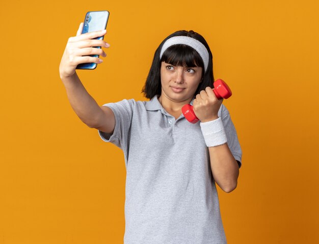 Chica joven fitness con diadema sosteniendo mancuernas haciendo selfie con smartphone mirando confundido parado sobre naranja