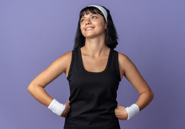 Chica joven fitness llevar diadema mirando hacia arriba sonriendo alegremente feliz y positivo de pie sobre fondo azul.