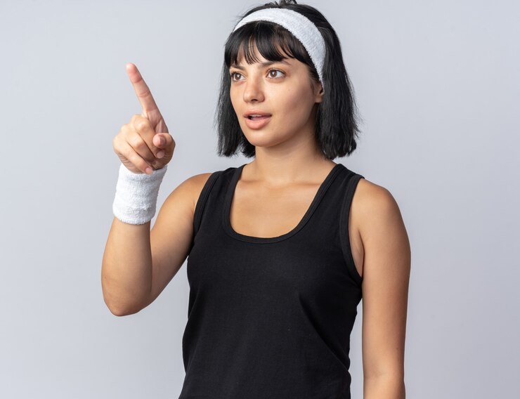 Foto gratuito chica joven fitness llevar diadema mirando a un lado intrigado apuntando con el dedo índice a algo de pie sobre fondo blanco.