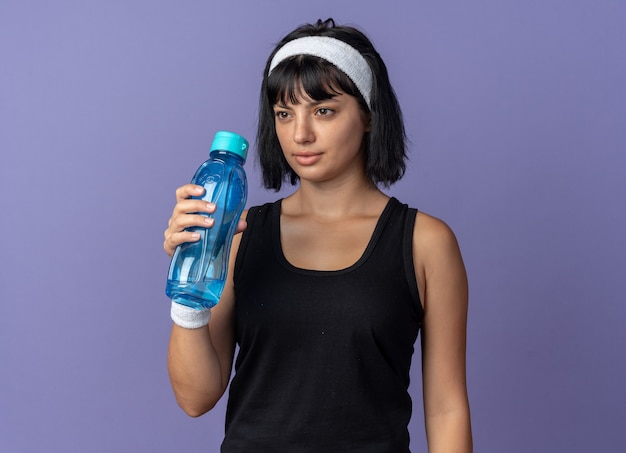 Chica joven fitness llevar diadema sosteniendo una botella de agua mirando a un lado con cara seria de pie sobre fondo azul.