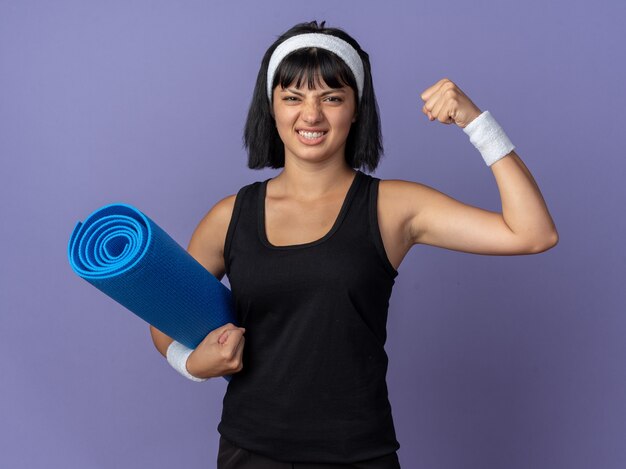 Chica joven fitness llevar diadema sosteniendo estera de yoga levantando el puño tenso y seguro de pie sobre fondo azul.