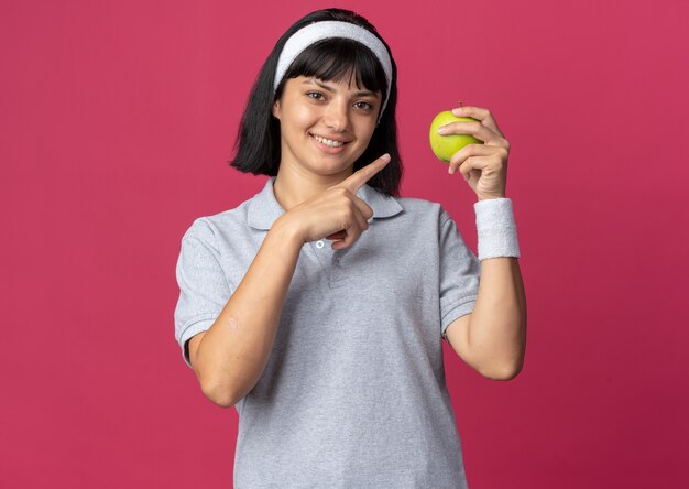 Chica joven fitness llevar diadema sosteniendo manzana verde apuntando con el dedo índice a Apple sonriendo alegremente de pie sobre fondo rosa