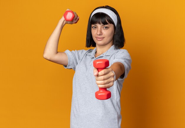 Chica joven fitness llevar diadema sosteniendo pesas haciendo ejercicios mirando confiados de pie sobre fondo naranja