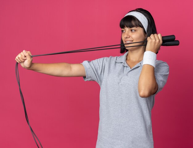 Chica joven fitness llevar diadema sosteniendo saltar la cuerda mirando a un lado con una sonrisa en la cara feliz de pie sobre fondo de color rosa