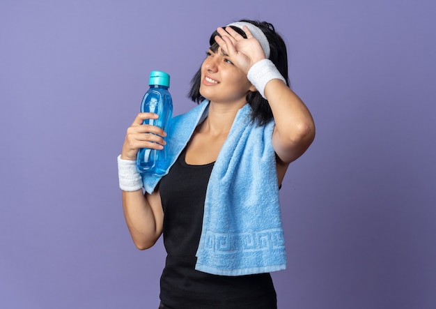 Chica joven fitness llevar diadema con una toalla alrededor del cuello sosteniendo una botella de agua con aspecto cansado y con exceso de trabajo de pie sobre fondo azul.