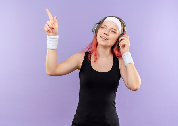 Chica joven fitness en ropa deportiva con auriculares en la cabeza sonriendo apuntando con el dedo hacia el lado de pie sobre la pared púrpura