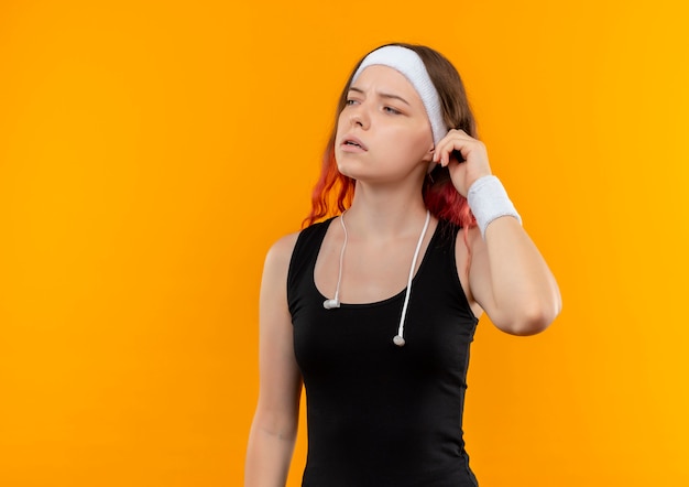 Chica joven fitness en ropa deportiva con auriculares mirando a un lado con expresión pensativa de pie sobre la pared naranja